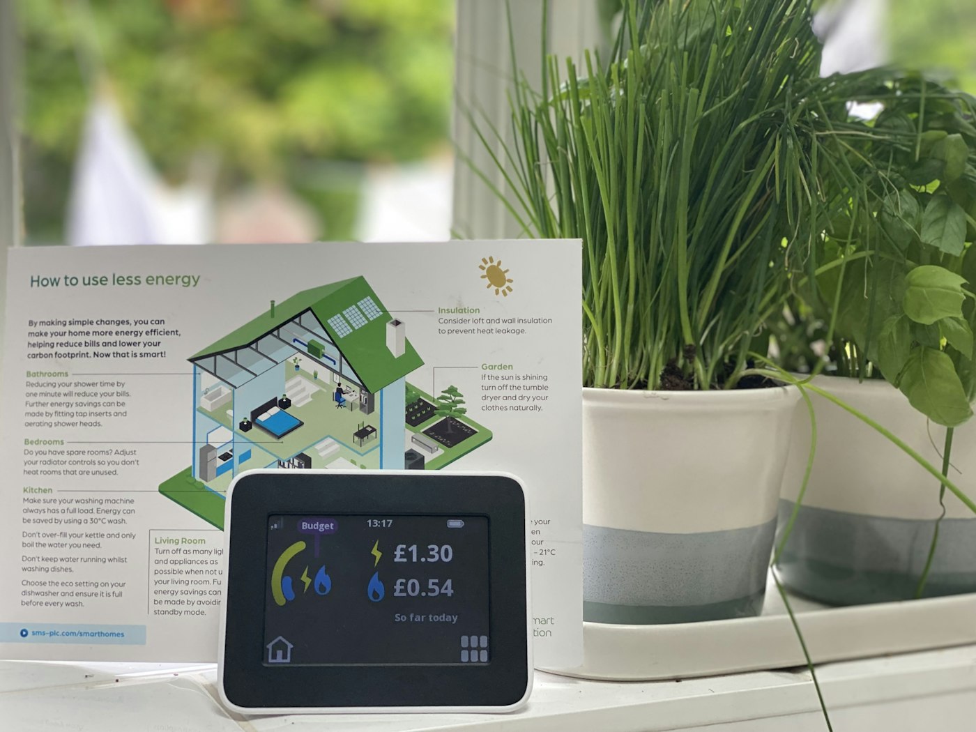 Smart meter dashboard showing electricity and gas energy consumption on kitchen window sill, next to herb plants. Laundry drying outside in background. 